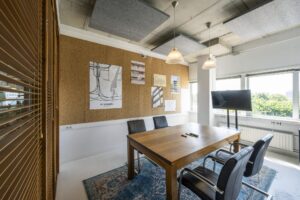 Vergaderruimte met houten tafel, vier zwarte stoelen, een prikbord met architectuurposters en een scherm bij het raam aan de Anthony Fokkerweg.